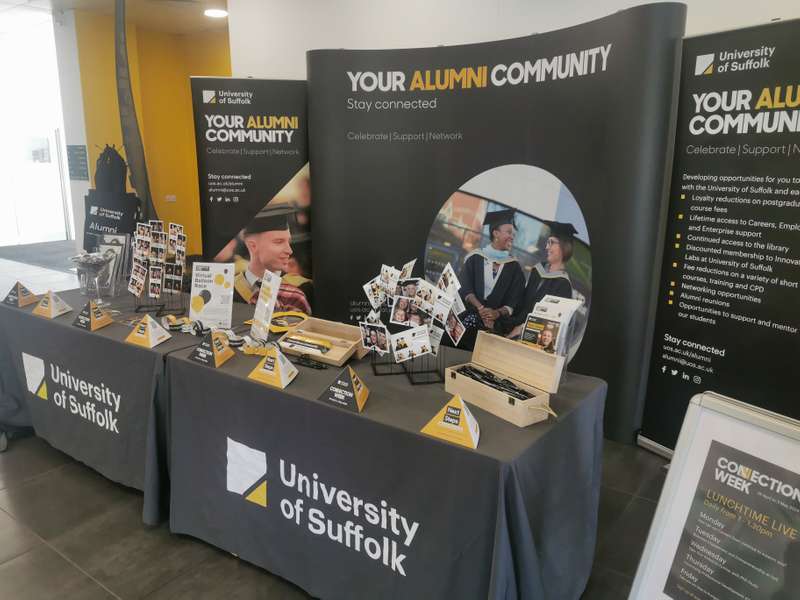 Promotional event area for the University of Suffolk alumni community, featuring several display tables with brochures, banners, and photographs.