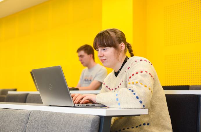 A student sitting in the lecture theatre typing at a laptop. Another student is visible in the row behind