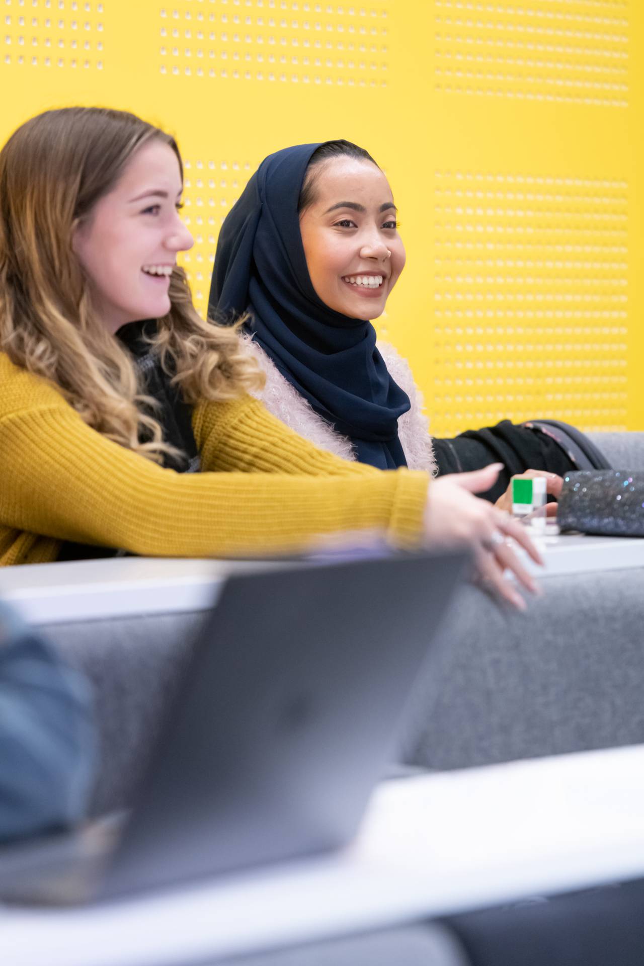 Two students sat in a lecture yellow background behind them