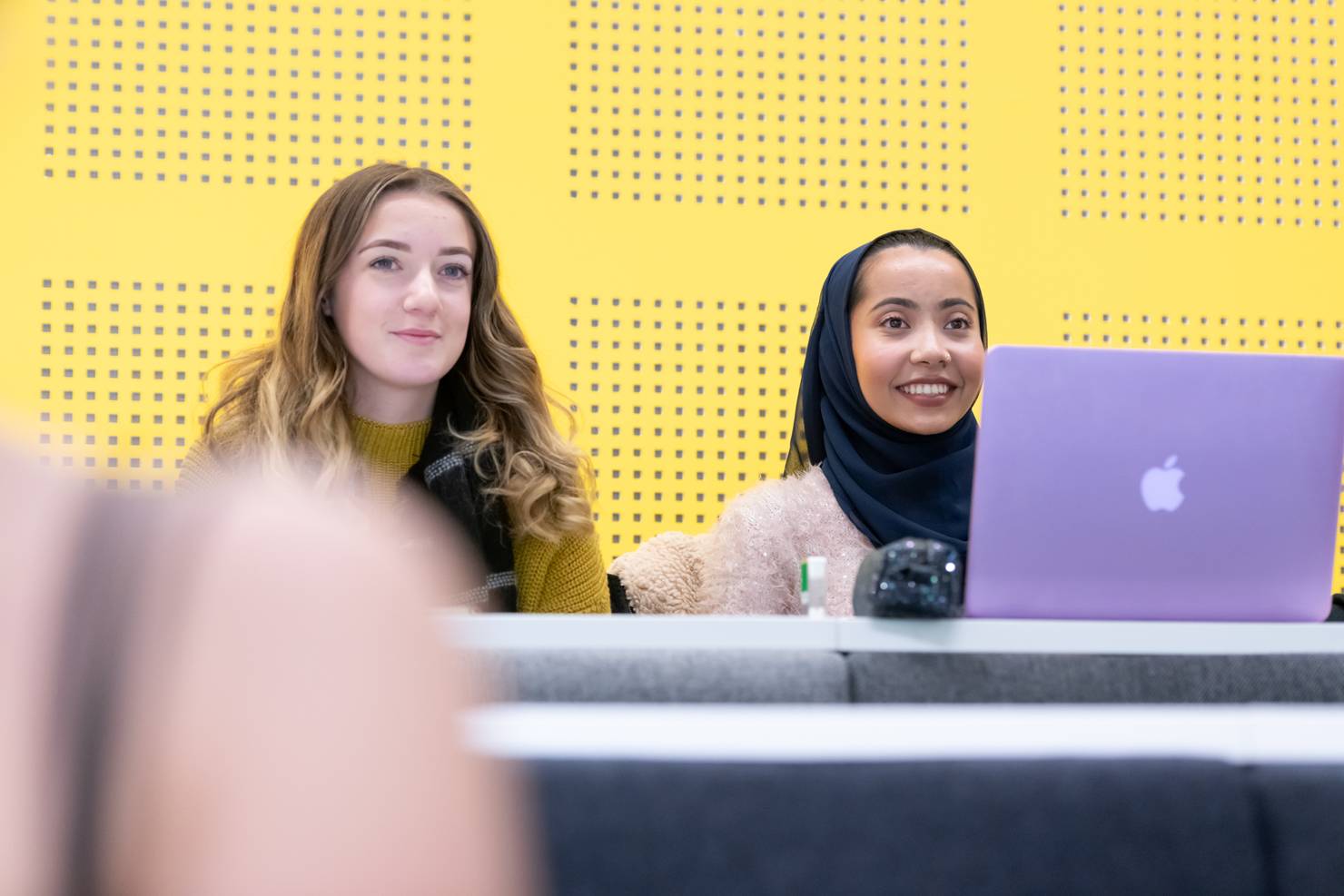 Two students sat in a lecture with a yellow wall behind them