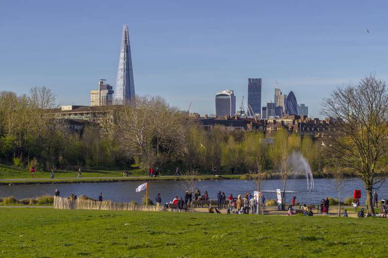 People gathered by the waters edge at Burgess park, lake behind them and the London skyline in the background, including the Shard, the Gherkin and the Fenchurch Building (walkie-talkie building)
