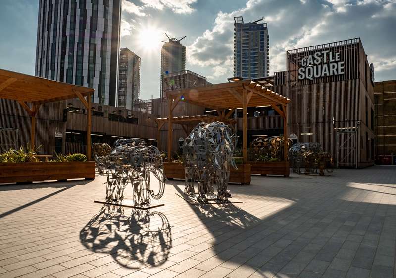 Lifesize elephant statues outdoors in the Castle Square market in London. Tall buildings in background with sunlight poking out behind.