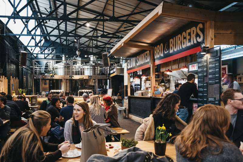 People sitting at tables eating in an indoor market, Mercato Metropolitano, BBQ and burger stand in background.