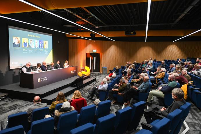 A public lecture happening in The Hold lecture theatre. Members of the public have filled the lecture seats while a desk and panel of five speakers are at the front of the lecture theatre
