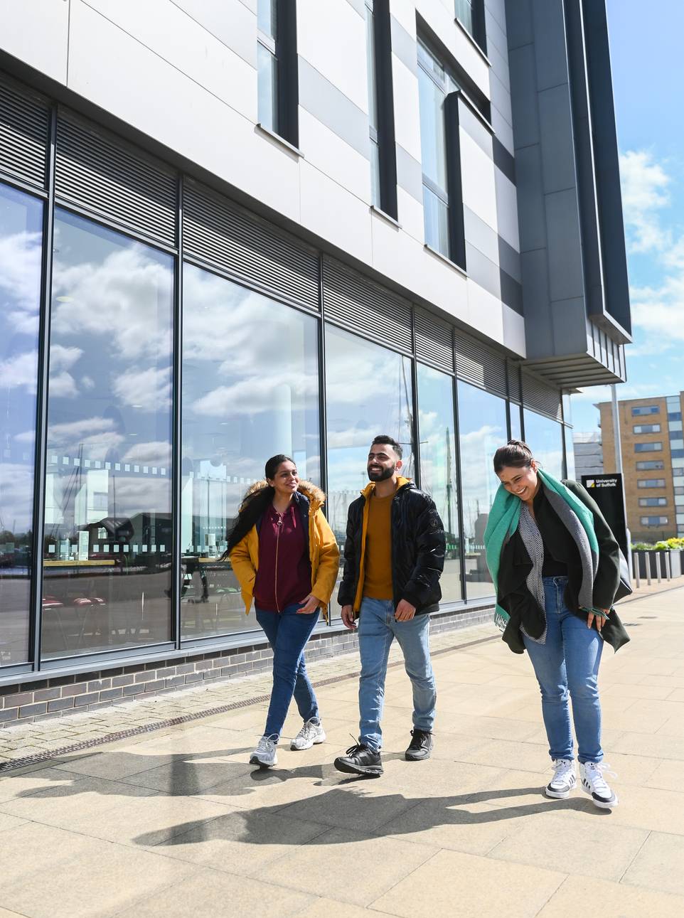 Three students walking past the glass windows of the waterfront building on a sunny day