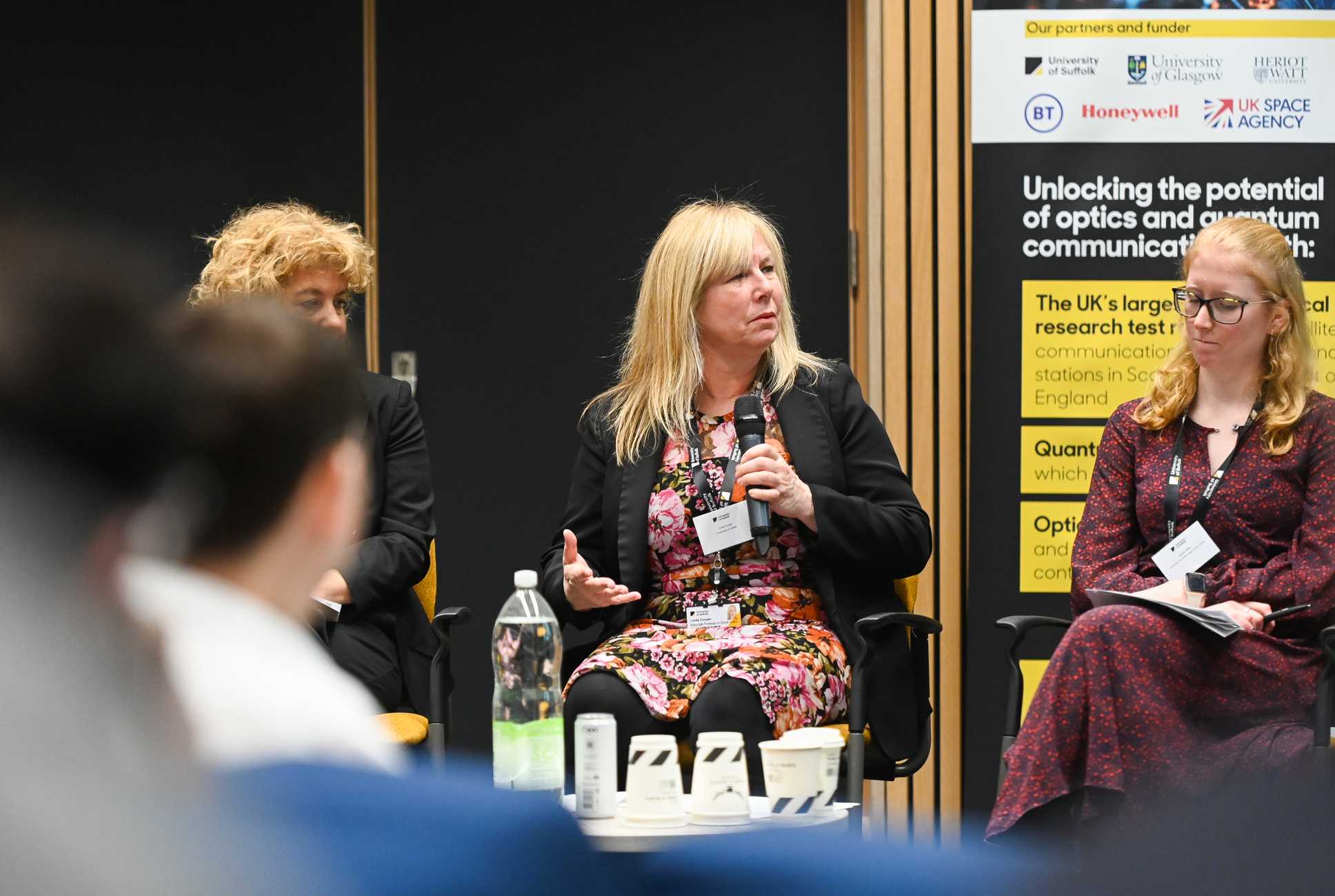 Three speakers sat in chairs, one with a microphone. A yellow and black banner sits behind the furthest speaker.